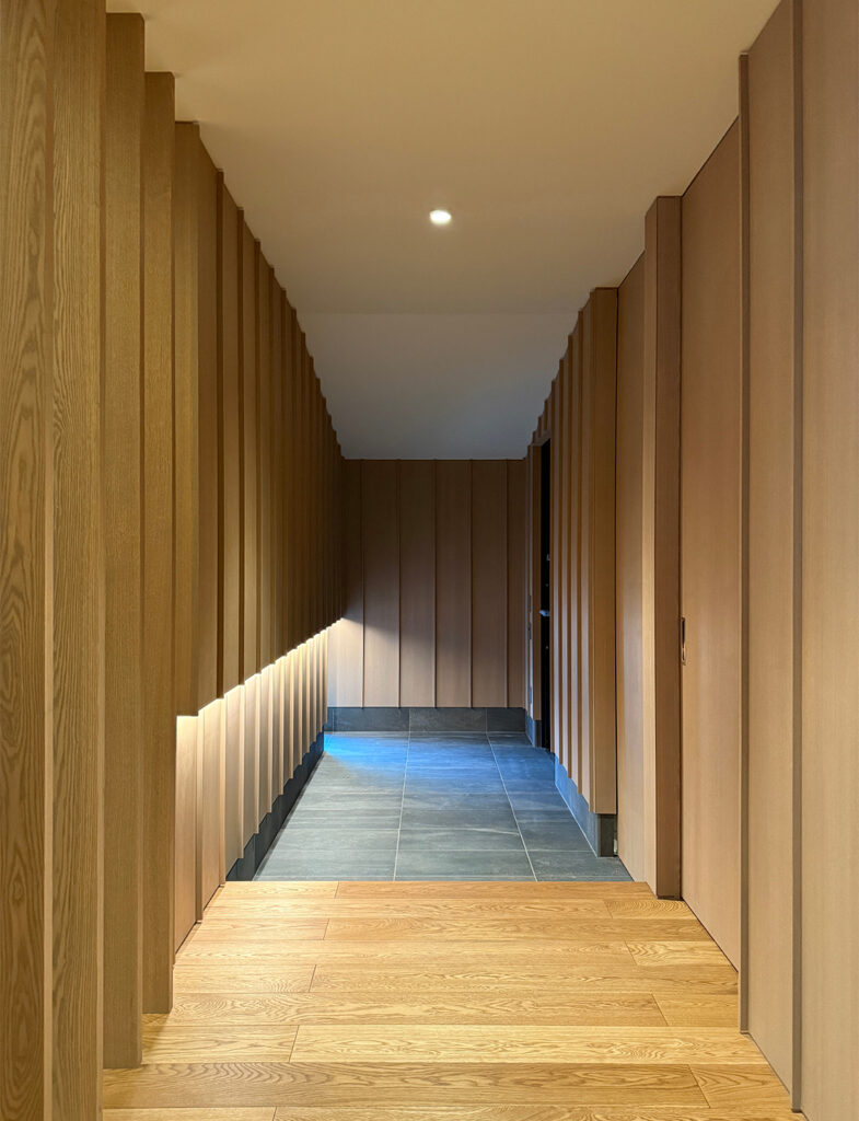 温かみのある木材と直線的なラインが印象的な、モダンで静謐な廊下の写真。 壁面には等間隔に配置された垂直な木製ルーバーが並び、左側の壁の中ほどには水平に伸びる間接照明が組み込まれています。床材は手前が明るい色の木目調フローリング、奥は落ち着いたグレーのタイルに切り替わっており、天井には小さなダウンライトが1つ灯っています。全体的に和モダンな雰囲気を感じさせるデザインです。