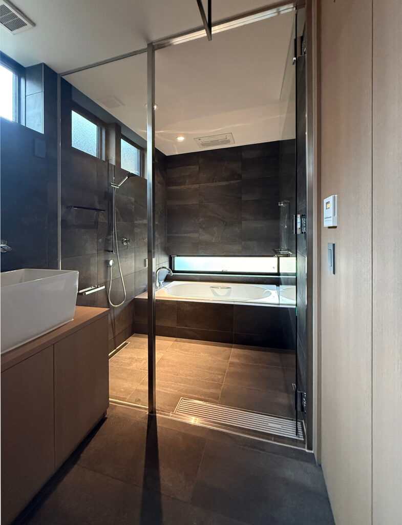 ガラス張りの仕切りが開放的な、高級感あふれるバスルームの風景。壁と床にはマットな質感のダークグレーの大理石調タイルが使われ、シックな印象を与えています。奥には白い浴槽が配置され、そのすぐ上の低い位置に設けられた横長の窓から自然光が差し込んでいます。手前には洗面台の一部が見え、ガラス戸のフレームや水栓のシルバーがアクセントとなった、和モダンでホテルライクな空間です。