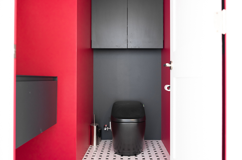 鮮やかな赤とマットな黒のコントラストが目を引く、モダンで大胆なトイレ空間。左右の壁面には濃い赤色の塗装が施され、正面の壁とタンクレストイレ本体、上部の収納棚はすべてマットブラックで統一されています。床には白と黒の小さなタイルが幾何学模様のように並び、白いドアとの対比が空間の個性を際立たせています。住宅全体の落ち着いたトーンの中に現れる、サプライズのようなデザイン空間です。