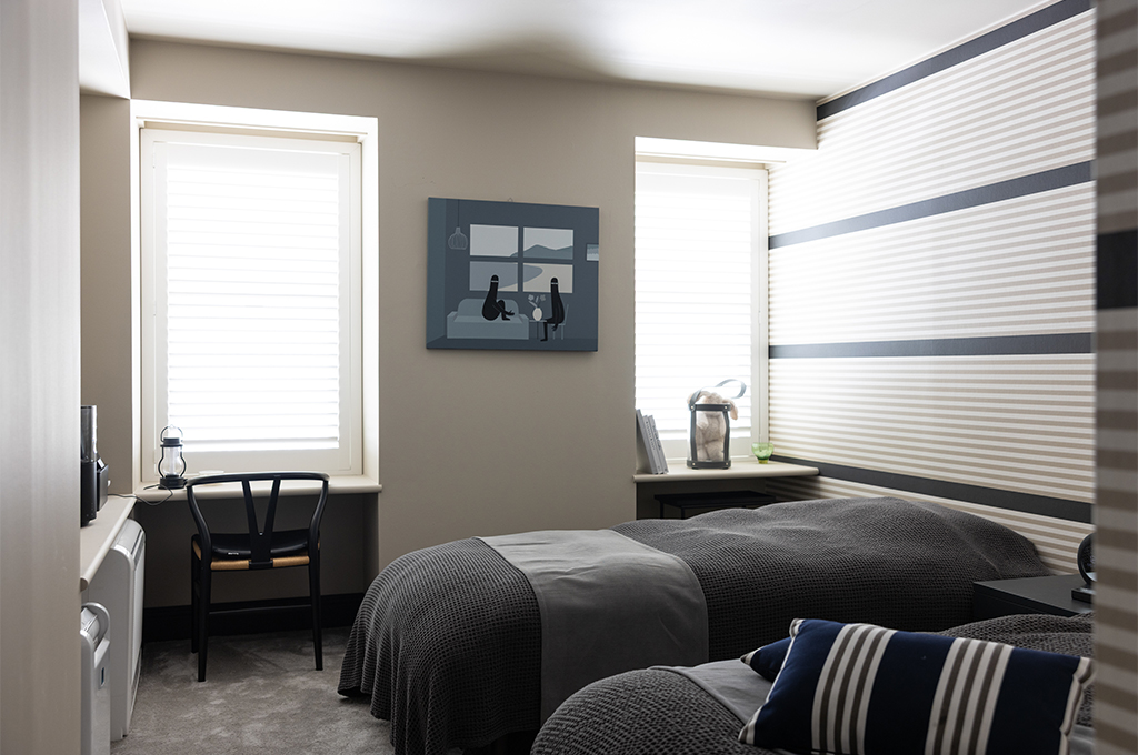 落ち着いたグレージュの壁面と、ボーダー柄のアクセントクロスが印象的なツインベッドルーム。窓辺には備え付けのデスクカウンターがあり、ハンス・J・ウェグナーの「Yチェア（CH24）」の黒が空間を引き締めています。ベッドにはグレーのワッフル織のリネンが掛けられ、壁にはリビングとお揃いのアートが飾られています。柔らかい自然光が差し込む、静かで整った心地よさを感じさせるプライベート空間です。