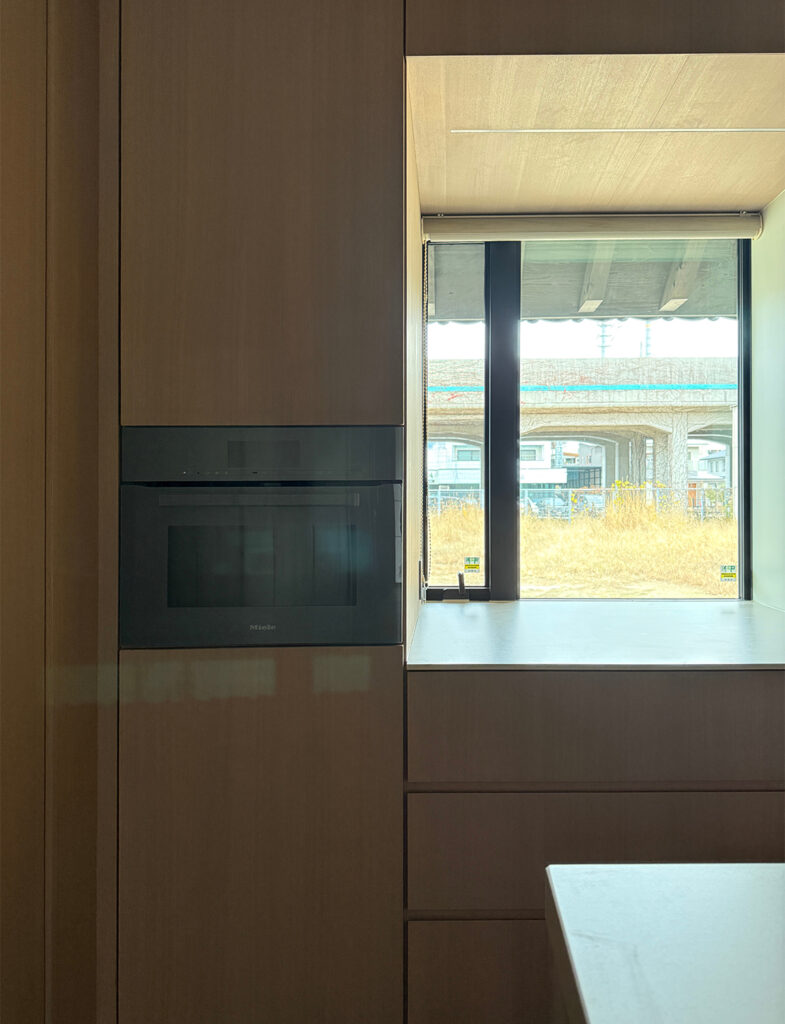 壁面の木製収納に美しく組み込まれた、Miele(ミーレ)製のビルトインオーブン。その隣にはスクエアな窓があり、ワークトップが窓辺まで伸びて機能的なスペースを作り出しています。窓の外にはのどかな風景が広がり、直線の構成が美しい和モダンの機能美を象徴するカットです。