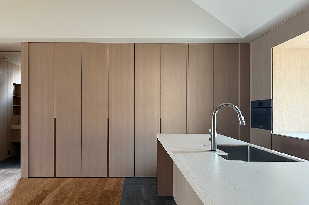 キッチン背面の大型収納とアイランドカウンターを捉えたカット。壁一面を占める収納扉は、継ぎ目を感じさせないミニマルなデザインで、温かみのある木目(またはSkimming Stoneと調和する色調)で統一されています。手前のアイランドカウンターは白い天板で、スタイリッシュな混合水栓とシンクが備わっています。床材は、フローリングからキッチンエリアのダークグレータイルへと切り替わっており、素材のコントラストが空間にリズムを与えています。