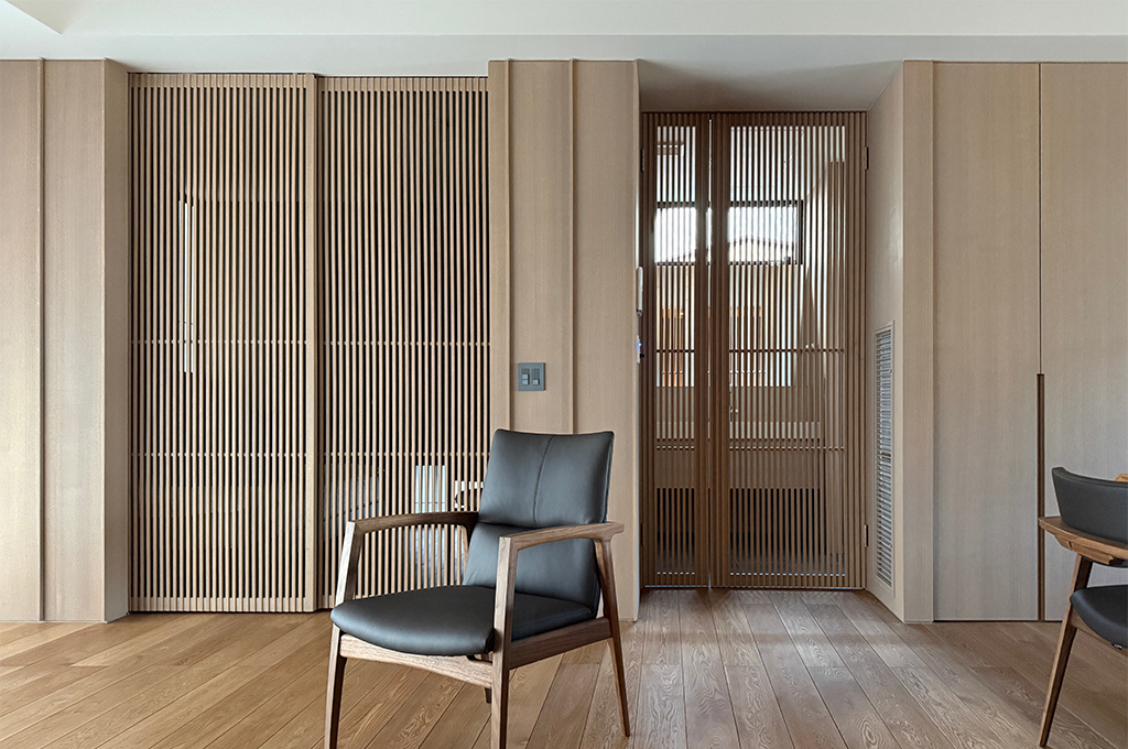 パターン1:詳細な説明 質感の良い黒いレザーと木製フレームのラウンジチェアを主役に据えた、リビングの一角。背景には、空間を緩やかに仕切る繊細な縦格子の引き戸と、壁一面のフラットな木製収納が並んでいます。格子の隙間からは奥の空間の光が漏れ、フローリングの木目と相まって、静かで知的な和モダンの雰囲気を醸し出しています。