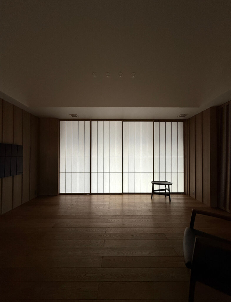 薄暗い部屋の奥に、光を透かす大きな障子戸が広がる静かな和モダンの空間。 正面の壁一面が4枚の障子戸になっており、外からの柔らかな光が室内を照らしています。障子の手前右側には小さな黒いサイドテーブルが置かれ、手前右端には椅子の背もたれの一部が見えます。床は落ち着いた色合いの幅広のフローリングで、左右の壁は垂直なラインが際立つ木目パネルで仕上げられています。天井には小さなダウンライトが並び、ミニマルで瞑想的な雰囲気を感じさせる写真です。