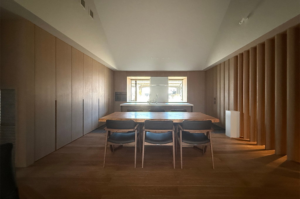 勾配天井(三角屋根のような形状)が開放的な、木のぬくもり溢れるモダンなダイニングキッチンの写真。 中央には重厚な木製の一枚板のようなダイニングテーブルがあり、黒いレザー張りの椅子が3脚並んでいます。その奥には白いカウンタートップのアイランドキッチンがあり、正面の窓からは外光が差し込んでいます。左側の壁は一面がフラットな木製収納扉になっており、右側の壁は等間隔に並んだ垂直な木製ルーバーのデザインになっています。天井は白く、照明を抑えた落ち着いたトーンの空間です。