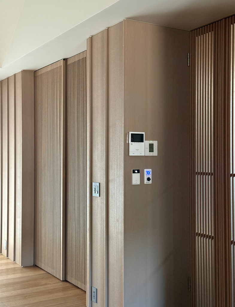 木目調の壁面にインターホンやスイッチ類が整然と配置された、機能的な室内の一角。 壁は垂直なラインの凹凸があるデザインで、中央のフラットな面にインターホンモニター、給湯器リモコン、照明スイッチ、調光ダイヤルが並んでいます。左奥には細い格子状の引き戸が見え、右側にも同様の木製ルーバーが設置されており、統一感のある和モダンな内装です。床は明るい色のフローリングで、壁の最下部にはコンセントプレートも配置されています。