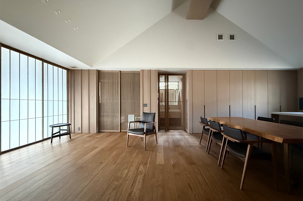 追加でアップロードいただいた広角のリビングダイニングの画像(6.jpg)についても、代替テキストを作成しました。これで空間全体の繋がりがより分かりやすくなりますね。
1枚目(リビングダイニングの全景 / 6.jpg)
パターン1:詳細な説明 勾配天井(三角屋根)による高い吹き抜けが印象的な、広々としたリビングダイニングの全景。左側には柔らかな光を通す大きな障子戸、中央には格子状の引き戸、右側にはフラットな木製収納扉と、異なる木の表情が壁面を構成しています。手前には木目調のフローリングが広がり、黒いレザーのダイニングチェアとテーブル、そして中央に一脚のラウンジチェアが置かれた、ミニマルで洗練された和モダンの大空間です。