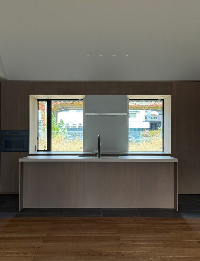 正面から捉えた、ミニマルで機能的なアイランドキッチンの風景。キッチン本体は壁面と同じく落ち着いた木目調で構成され、天板には清潔感のある白い素材が使われています。背後の壁には大きな開口部があり、シンクの正面に位置する窓からは外の風景と自然光が取り込まれています。床は手前が木目フローリング、キッチン足元がダークグレーのタイルに切り替わっており、視覚的なゾーニングがなされています。