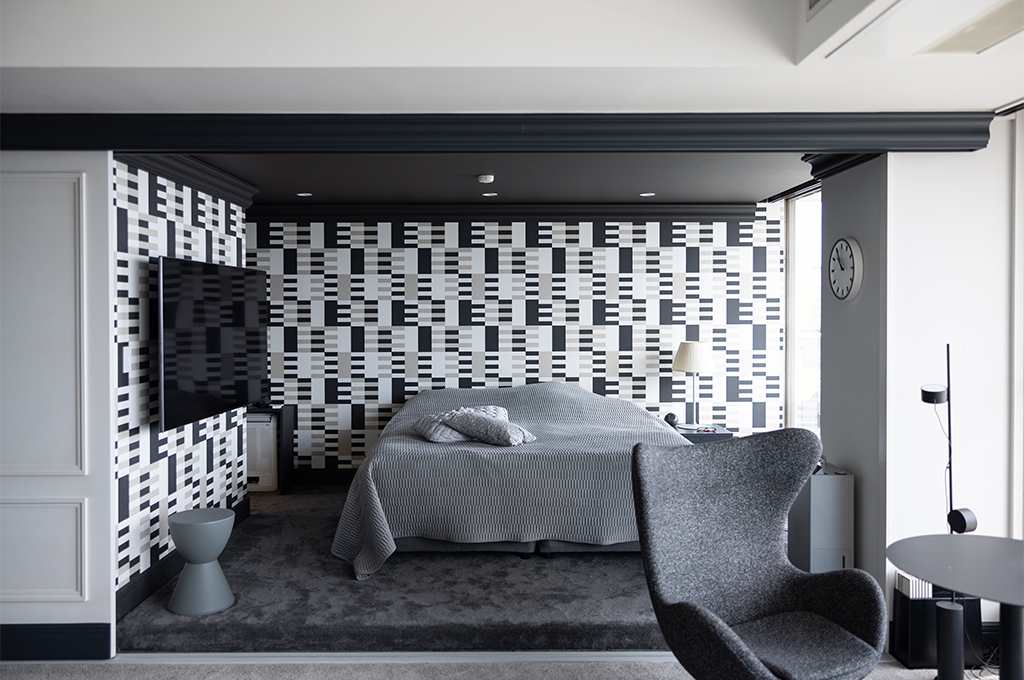 モノトーンの幾何学模様の壁紙が目を引く、アーティスティックな寝室。ロータイプの広いダブルベッドにはグレーのリネンが掛けられ、天井に近い梁の部分は黒いモールディングで縁取られています。手前にはグレーのアルネ・ヤコブセン「エッグチェア」とサイドテーブルが配され、奥には壁掛けテレビが設置されています。寝室でありながら、ラウンジのような高級感漂うデザインです。