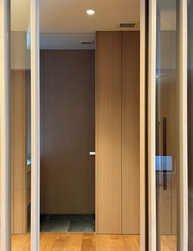 木製の縦格子引き戸を開けた先に見える、室内動線のカット。正面にはフラットな仕上げの木製建具(ドア)があり、手前の明るいフローリングから、奥のグレーのタイル床へと繋がっています。天井のダウンライトが一点、木の壁面を優しく照らし、直線的なラインが重なり合うことで、和モダンな建築特有の「隙間の美」を表現しています。