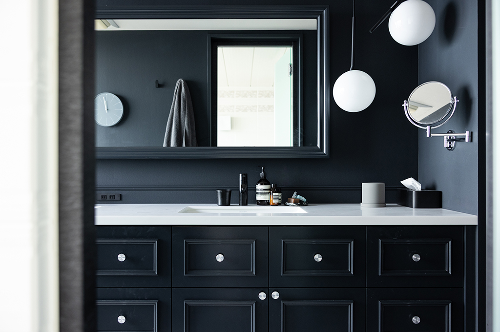 ブラックで統一された、ホテルライクでドラマチックな洗面スペース。壁面、クラシックなモールディングが施されたキャビネット、鏡のフレームに至るまで深い黒で仕上げられ、白いワークトップとのコントラストが際立っています。鏡の上には丸いフォルムの白いペンダントライトが浮遊するように配され、Aesop（イソップ）のボトルやシルバーの水栓が上質なアクセントを添えています。一貫した美学が感じられる、洗練された空間です。