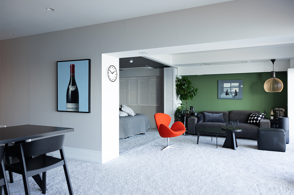 白を基調とした開放的な空間に、印象的な色彩が散りばめられたモダンなリビングルーム。手前のダイニングエリアには黒いテーブルと椅子が配され、壁面には大きなワインボトルのアートが飾られています。奥のリビングスペースには、鮮やかなオレンジ色のアルネ・ヤコブセン「スワンチェア」が鎮座し、グレーのソファの背後にある壁一面には、落ち着いたグリーンの塗装が施されています。床には一面に白いカーペットが敷き詰められ、異なるエリアがゆるやかに繋がりつつも、それぞれの「色」によって明確な個性が与えられた洗練された空間です。