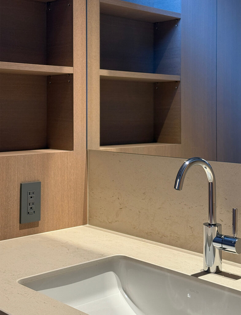 洗練されたデザインの洗面コーナーのアップ。ベージュトーンの石目調カウンターに、スクエア型の白い洗面ボウルと、曲線が美しいクローム仕上げの混合水栓が設置されています。正面には大きな鏡があり、木製のオープンシェルフが映り込んでいます。左側の木目壁には、空間のトーンに合わせたグレーのコンセントプレートが配置され、細部まで色彩設計が徹底された清潔感のある空間です。