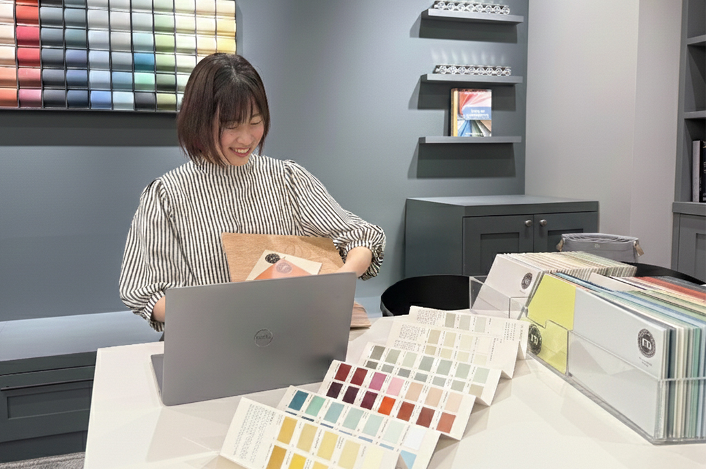 「オンラインカラーコンサルタンシーの様子。画面越しに大きな色サンプルを示しながら、コンサルタントが丁寧に説明しているシーン。お客様の手元には事前に送付されたカラーチャートがあり、双方で色を確認し合いながら進めている。