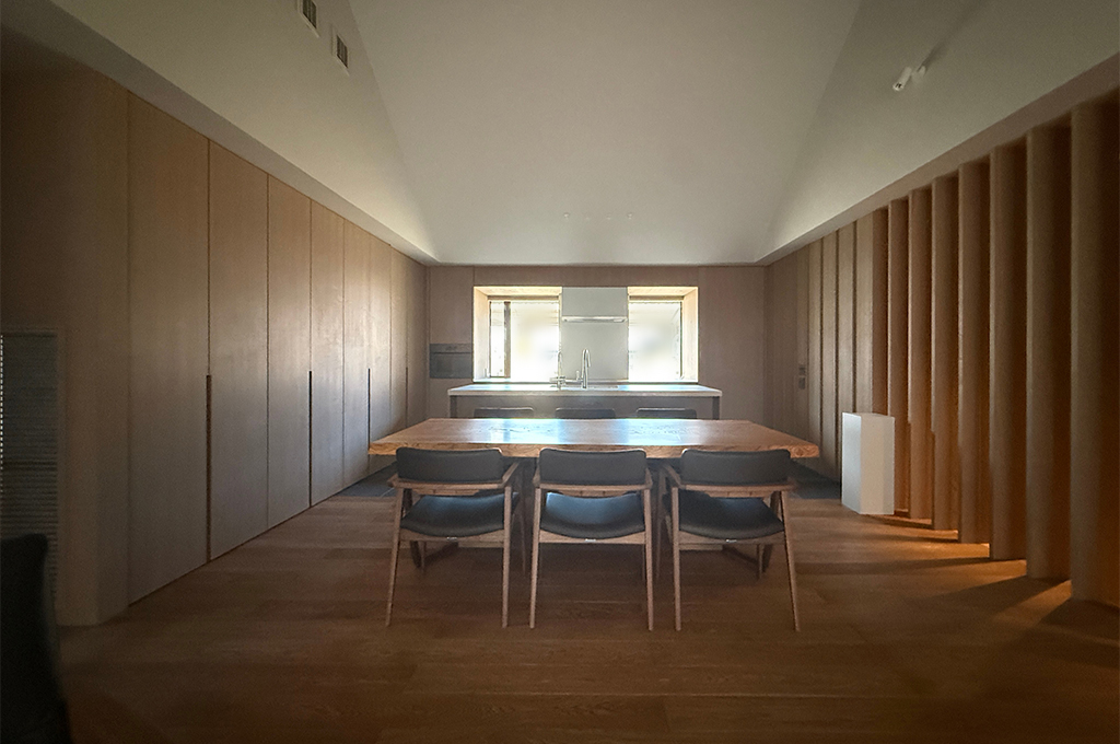 勾配天井（三角屋根のような形状）が開放的な、木のぬくもり溢れるモダンなダイニングキッチンの写真。 中央には重厚な木製の一枚板のようなダイニングテーブルがあり、黒いレザー張りの椅子が3脚並んでいます。その奥には白いカウンタートップのアイランドキッチンがあり、正面の窓からは外光が差し込んでいます。左側の壁は一面がフラットな木製収納扉になっており、右側の壁は等間隔に並んだ垂直な木製ルーバーのデザインになっています。天井は白く、照明を抑えた落ち着いたトーンの空間です。