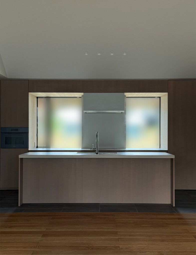 正面から捉えた、ミニマルで機能的なアイランドキッチンの風景。キッチン本体は壁面と同じく落ち着いた木目調で構成され、天板には清潔感のある白い素材が使われています。背後の壁には大きな開口部があり、シンクの正面に位置する窓からは外の風景と自然光が取り込まれています。床は手前が木目フローリング、キッチン足元がダークグレーのタイルに切り替わっており、視覚的なゾーニングがなされています。