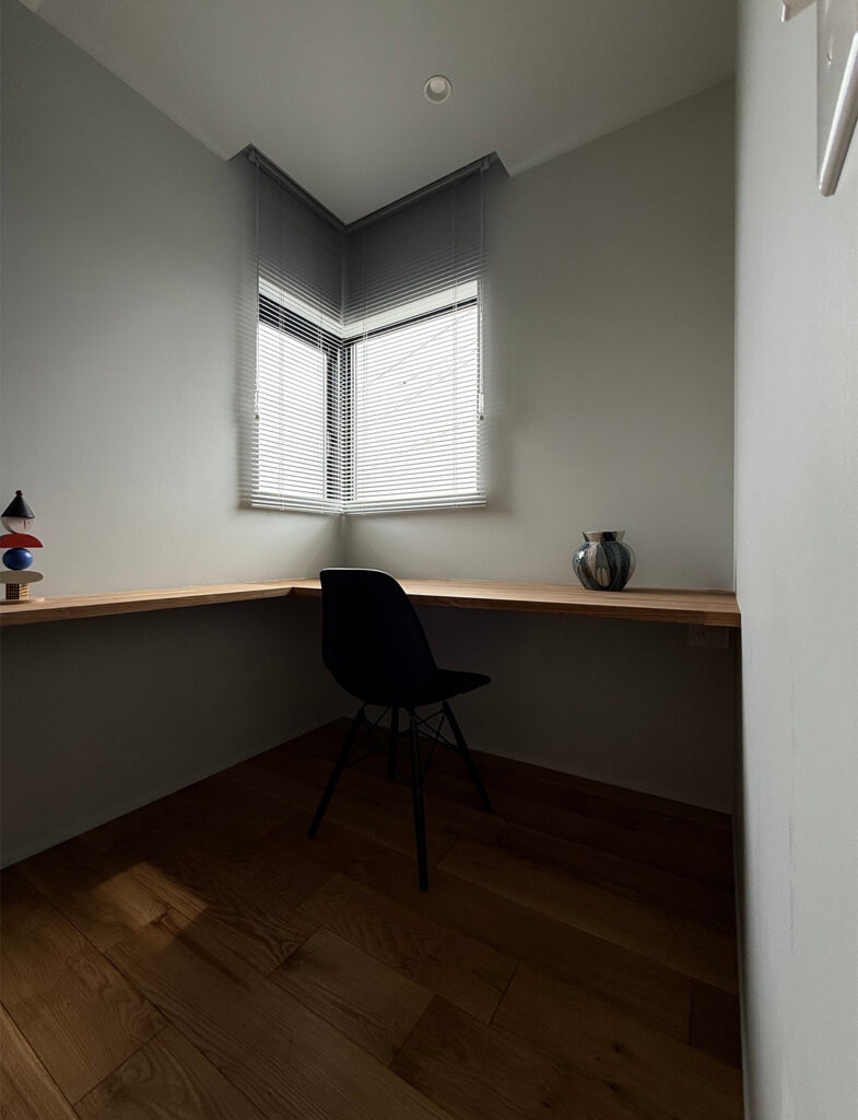 Farrow&Ballのペイントが光を柔らかく捉える、静謐な書斎コーナー。L字型のウッドデスクと黒のシェルチェアが、グレー系の壁面と美しくコントラストを描きます。ブラインドから漏れる光が、上質な空間の質感を引き立てるミニマルなインテリアです。