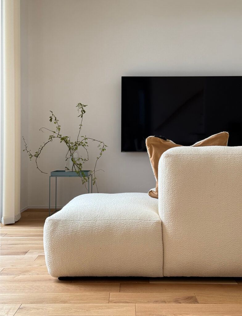 繊細なニュートラルカラーが、柔らかな素材感のソファと調和するモダンリビング。上質なペイント壁が木の温もりを引き立て、ミニマルながらも温かみのある北欧インテリアを実現しています。心地よい光が包む洗練された暮らしの風景。