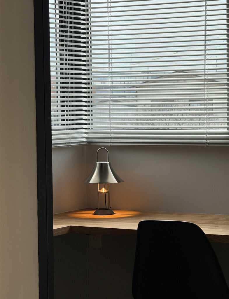 Farrow&Ballのマットな質感が際立つ、洗練された書斎のコーナー。ブラインド越しの柔らかな光と、デスクランプの温かな灯りが壁面の絶妙な色調を引き立てます。木目調のデスクとモダンな照明が調和する、上質なワークスペースの施工例です。
