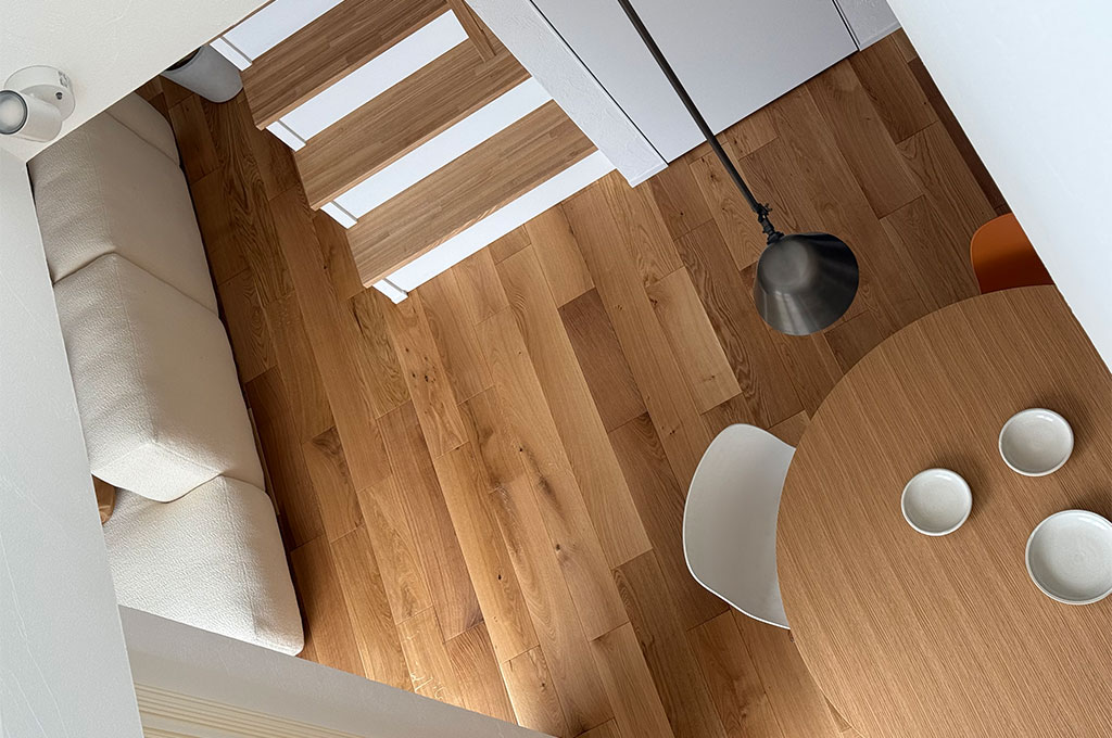 Farrow&Ballのマットな質感のペイント壁が、木の温もりあふれるリビングを俯瞰で包み込む。吹き抜けから見下ろすオーク材のフローリング、白いソファ、円形テーブルが織りなす、ミニマルで上質なナチュラルモダンスタイル。