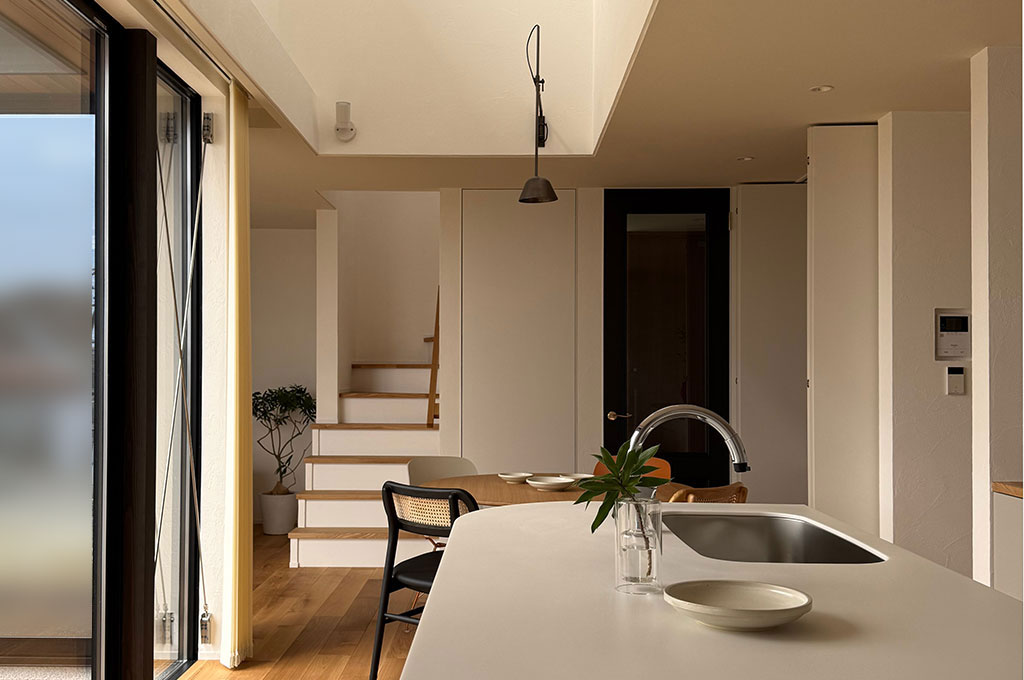 Farrow&Ballの柔らかなニュートラルカラーが開放的な吹き抜けを彩るキッチンダイニング。自然光が塗装壁に優しく反射し、モダンなアイランドキッチンや階段の木目と美しく調和する、心地よい静寂を感じさせる北欧モダンな住空間。