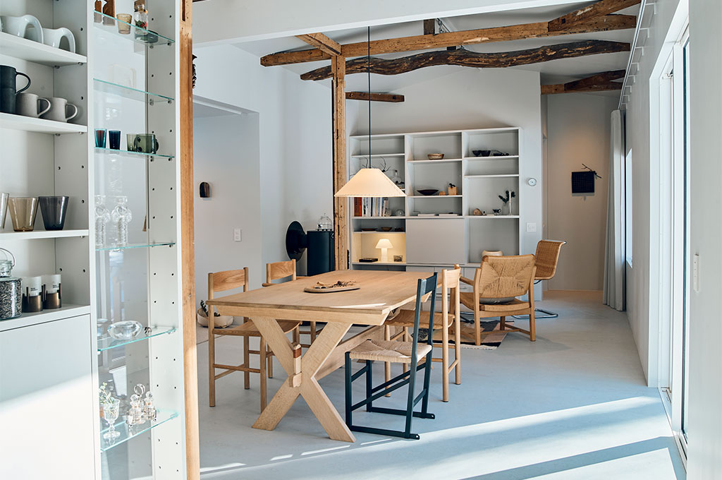 白を基調としたアトリエのダイニング。北欧のヴィンテージ家具を思わせる木製のダイニングテーブルとペーパーコードのチェアが、空間に温もりを与えている。左手の白いシェルフにはガラス器やオブジェが美しく整列し、背後の古材の梁が新旧の素材の対比を際立たせる。光が差し込む中で、機能性と美意識が同居したスタイリングの真骨頂。