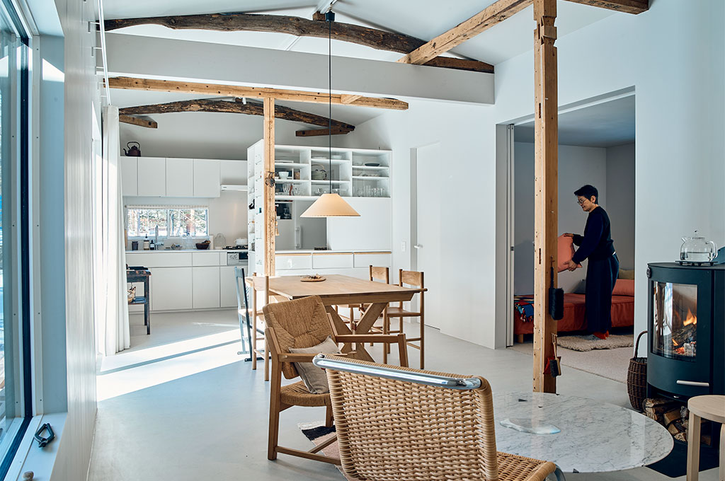 インテリアスタイリスト・黒田美津子さんのアトリエ内観。自然光が惜しみなく注ぐ真っ白な空間に、力強い古材の梁がコントラストを描く。ミニマルなキッチン、温かみのある北欧デザインの木製家具、そして生活に温もりを添える薪ストーブが配され、光と色が調和したスタイリッシュかつ心地よい居住空間を体現している。