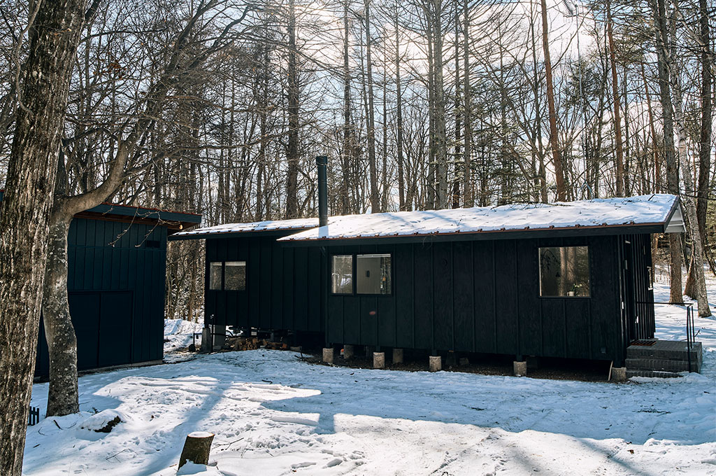 雪に覆われた静かな林間に佇む、黒い外壁のアトリエ。ミニマルな平屋建築に、光を反射する広々としたウッドデッキと大きなガラス扉が配され、自然と調和するモダンな空間づくりを象徴している。