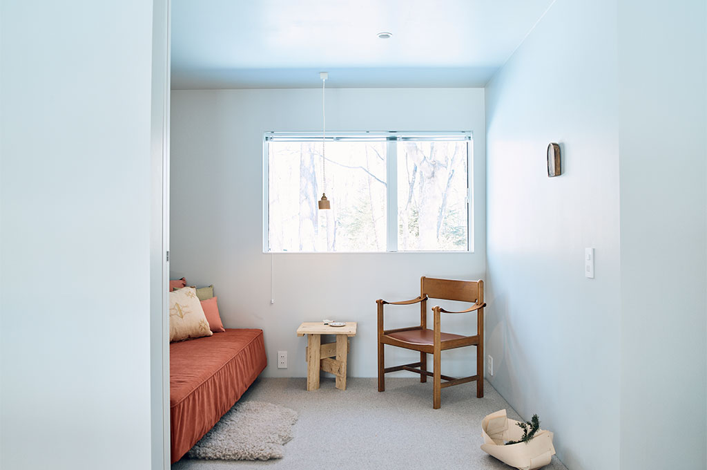 大きな窓から自然光が差し込む白い部屋のレイアウト。左手には暖色系のカバーがかかったデイベッドと複数のクッション、手前にはシープスキンのラグ。中央にはラフな質感の木製サイドテーブル、右手にはレザー座面の木製アームチェア（サファリチェア）が配置されている。窓の外には裸木が広がり、天井からは繊細なペンダントライトが吊るされている。