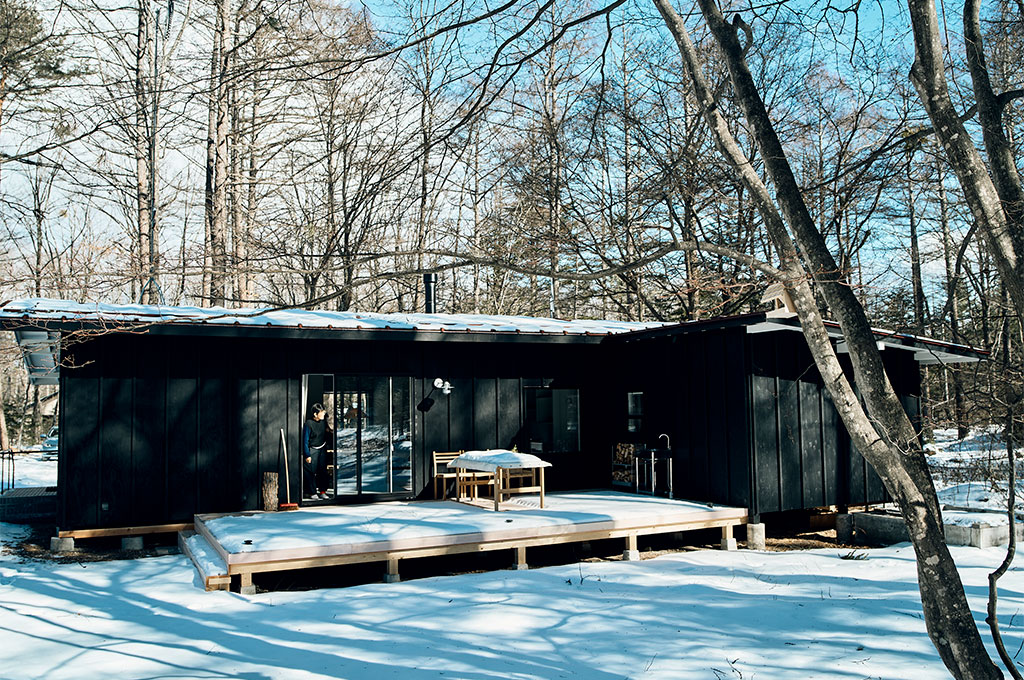 雪に覆われた静かな林間に佇む、黒い外壁のアトリエ。ミニマルな平屋建築に、光を反射する広々としたウッドデッキと大きなガラス扉が配され、自然と調和するモダンな空間づくりを象徴している。