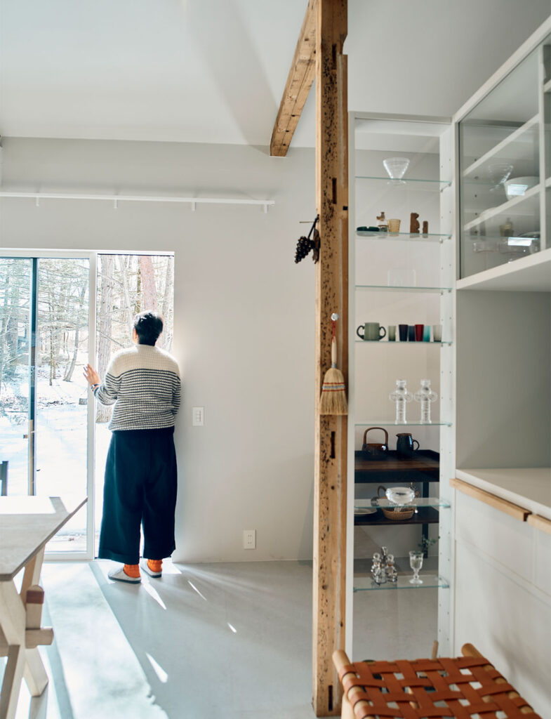 白を基調としたアトリエのダイニング。北欧のヴィンテージ家具を思わせる木製のダイニングテーブルとペーパーコードのチェアが、空間に温もりを与えている。左手の白いシェルフにはガラス器やオブジェが美しく整列し、背後の古材の梁が新旧の素材の対比を際立たせる。光が差し込む中で、機能性と美意識が同居したスタイリングの真骨頂。