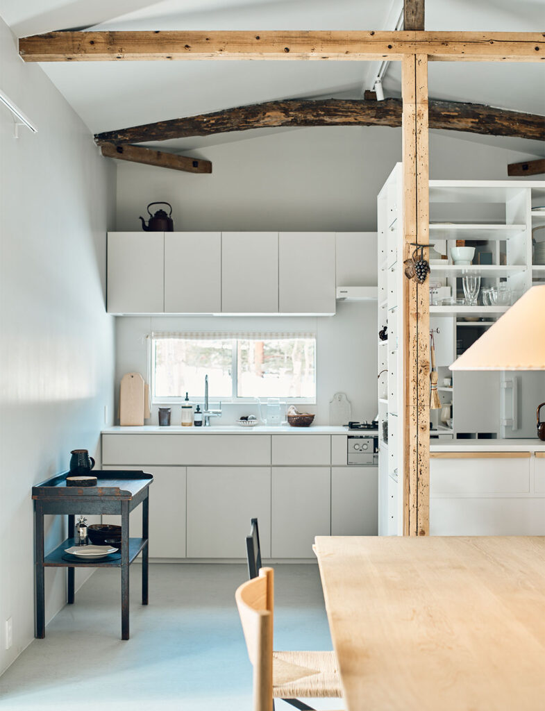 白を基調としたアトリエのダイニング。北欧のヴィンテージ家具を思わせる木製のダイニングテーブルとペーパーコードのチェアが、空間に温もりを与えている。左手の白いシェルフにはガラス器やオブジェが美しく整列し、背後の古材の梁が新旧の素材の対比を際立たせる。光が差し込む中で、機能性と美意識が同居したスタイリングの真骨頂。