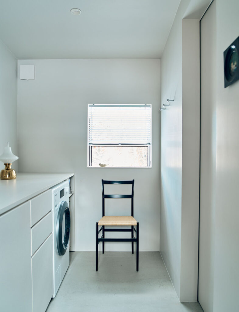 白で統一された洗練されたサニタリースペース。ミーレ（Miele）の洗濯機が組み込まれたミニマルなカウンターには、真鍮の輝きを放つヴィンテージランプや、季節を添えるグリーンの枝が飾られ、実用的な空間をアートのように演出している。木枠の鏡が白壁に温もりを添え、細部にまで黒田美津子さんの美意識が息づく。