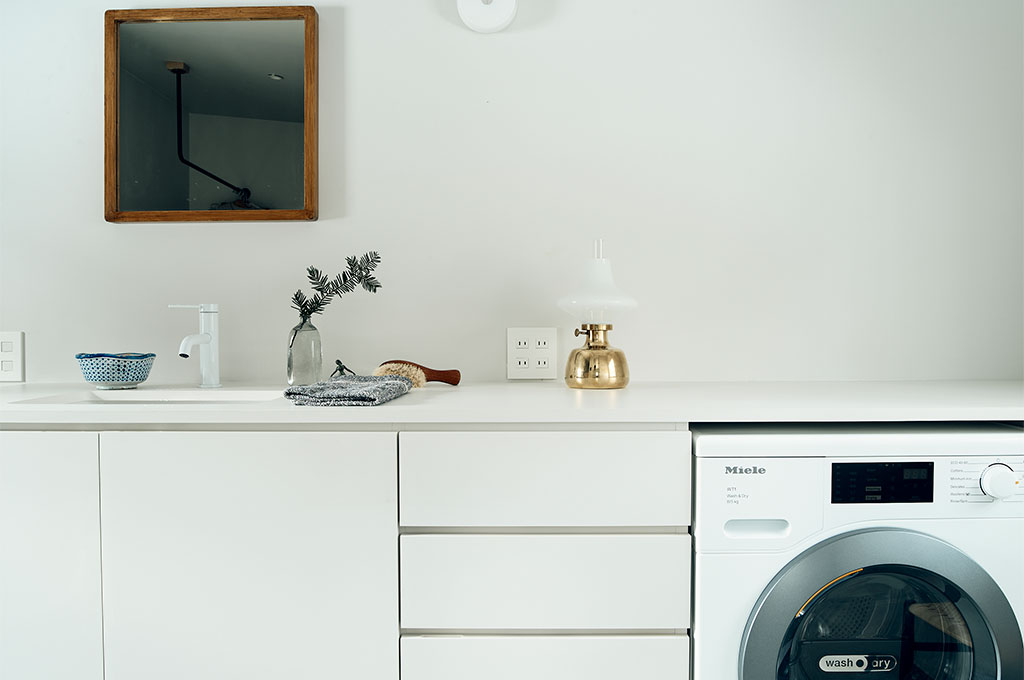 白で統一された洗練されたサニタリースペース。ミーレ（Miele）の洗濯機が組み込まれたミニマルなカウンターには、真鍮の輝きを放つヴィンテージランプや、季節を添えるグリーンの枝が飾られ、実用的な空間をアートのように演出している。木枠の鏡が白壁に温もりを添え、細部にまで黒田美津子さんの美意識が息づく。