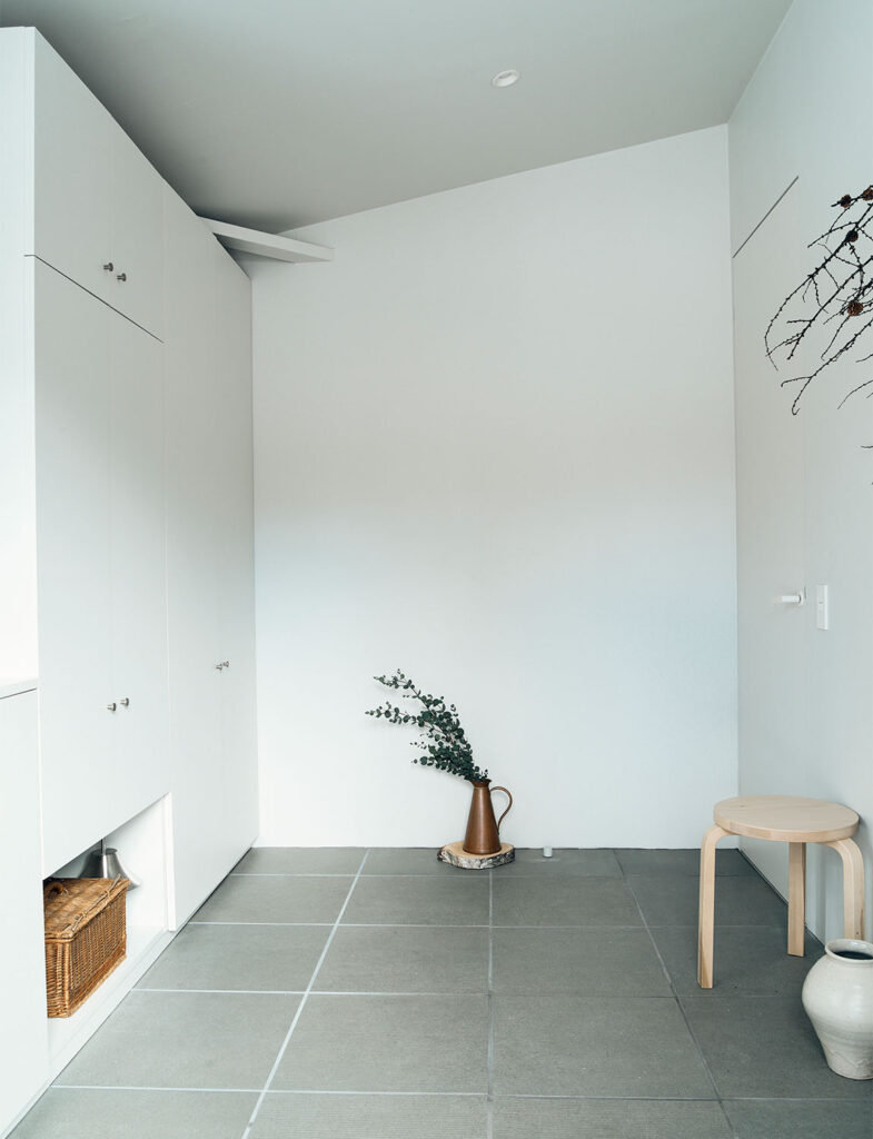 インテリアスタイリスト・黒田美津子さんのアトリエのエントランスホール。グレーのタイル床と白い壁面が織りなすミニマルな空間に、あえて大きな「余白」を残すことで、静謐な空気を生み出している。床に直置きされたブロンズ色のジャグとグリーンの枝、そしてアルヴァ・アアルトのデザインを彷彿とさせるスツールが、ギャラリーのような凛とした佇まいを添えている。