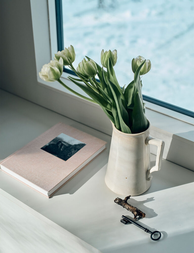 インテリアスタイリスト・黒田美津子さんのアトリエ窓辺を彩る静物。雪景色の青白い光を背景に、淡いピンクの装丁本、瑞々しい白いチューリップを生けた陶器のピッチャー、そして古びた鍵が並ぶ。暖色と寒色が繊細に響き合い、静かな冬の日の物語を予感させる、色彩と質感のプロフェッショナルなスタイリング。