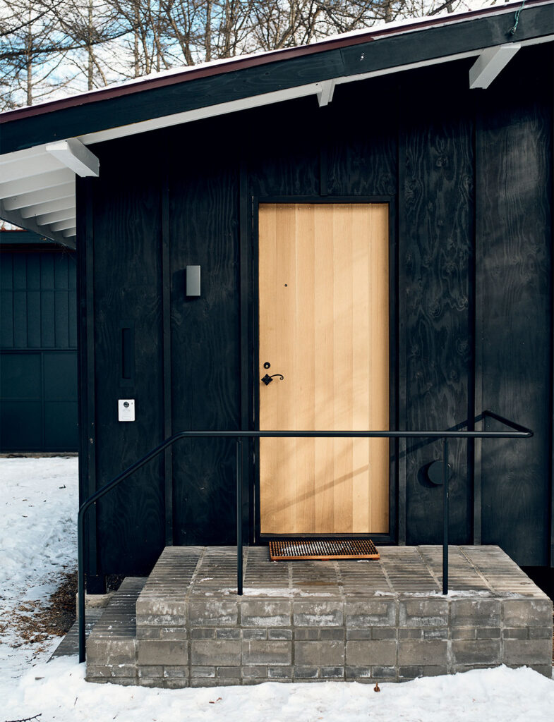 インテリアスタイリスト・黒田美津子さんのアトリエ玄関。焼き杉を思わせるマットな黒の木外壁に、白木の明るい玄関ドアが鮮やかなコントラストを描き出している。細身の黒いアイアン手すりと、質感豊かなレンガの階段が、ミニマルな建築にリズムと温もりを添える。厳かな冬の森に溶け込みながら、訪れる人を温かく迎え入れる「光と影」の象徴的なデザイン。