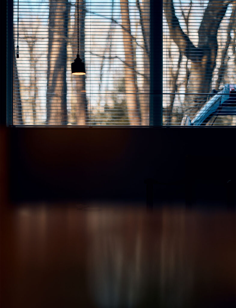 インテリアスタイリスト・黒田美津子さんのアトリエに訪れる、静かな夕暮れのひととき。ブラインドの隙間から漏れる夕陽が、冬の木立を背景に美しいストライプの光を室内へと運び、一灯のペンダントライトが闇の中に温かな居場所を作り出している。光と影が交差する瞬間の美しさを捉えた、空間の情緒を深く描き出すスタイリング。