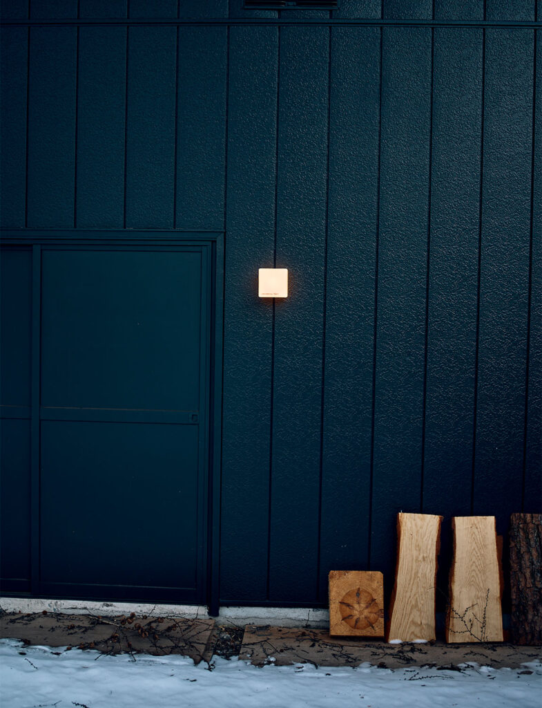 インテリアスタイリスト・黒田美津子さんのアトリエ、夜の表情。深い闇に溶け込む黒い縦羽目板の外壁に、正方形のブラケットライトが一灯、温かなオレンジ色の光を放っている。足元に立てかけられた薪の切り出しは、屋内の薪ストーブの温もりを予感させ、ミニマルな外観の中に「人の営み」を灯す、静謐でグラフィカルなスタイリング。