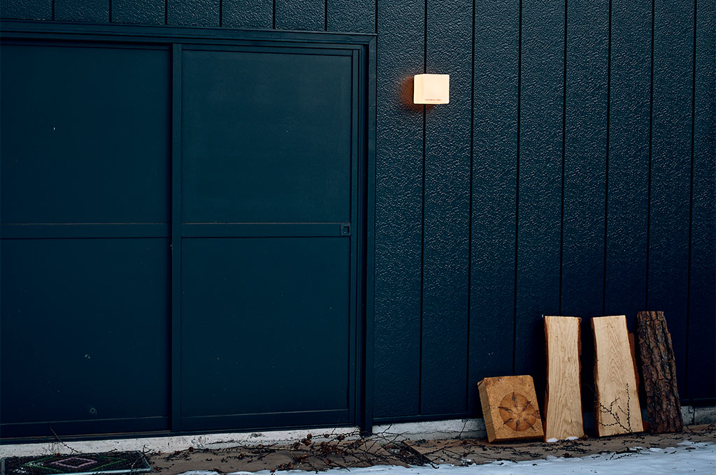 オフブラックの外壁に、一灯のブラケットライトが温かな光を投げかけるアトリエのディテール。壁面には質感の異なる木材の端材や耳付き板が立てかけられ、無機質な建材と有機的な自然素材が美しく共生している。静寂な夜の訪れを予感させる、光と色の繊細な構成。