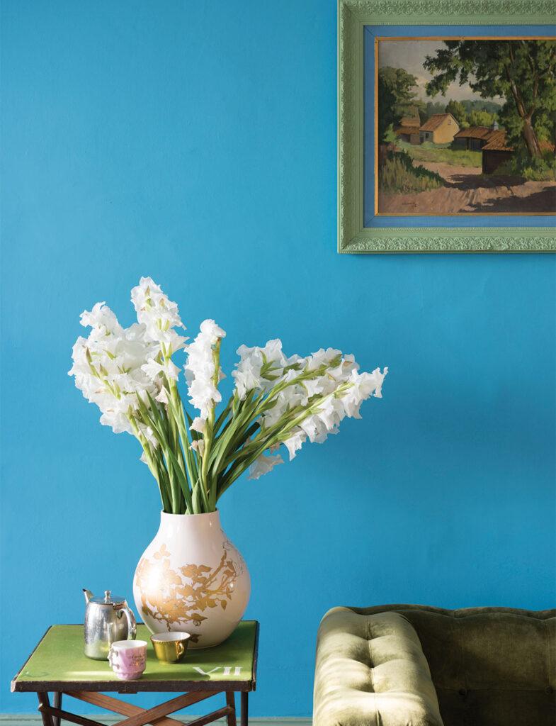 Farrow & Ballの鮮やかで清々しい「St Giles Blue」の壁が印象的なインテリア。オリーブグリーンのソファや白い花とのコントラストが美しく、クラシックな絵画が映える、遊び心溢れるエレガントな空間デザインです。