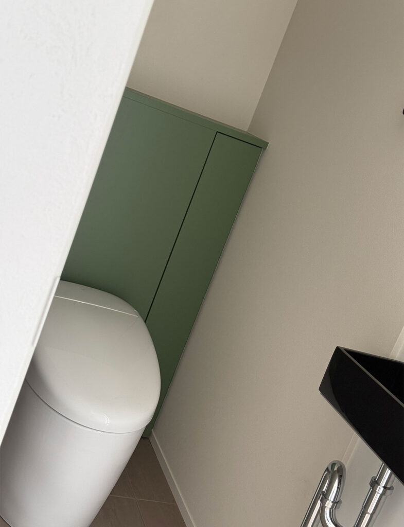 Farrow&Ballの深みのあるセージグリーン「Calke Green No.34」で塗装されたトイレ収納。落ち着いたベージュの壁や黒い手洗器と美しく調和し、コンパクトな空間に洗練されたアクセントを加えるレストルームです。