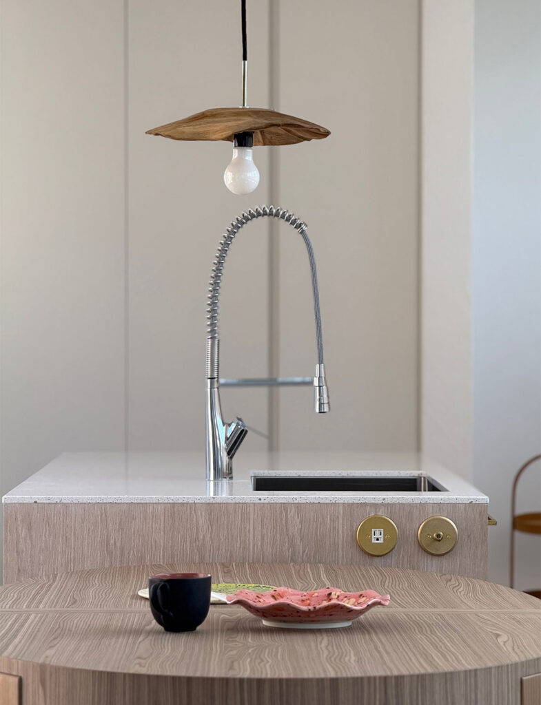 スタイリッシュな水栓が映えるアイランドキッチン。手前の木製ラウンドテーブルに置かれた器や、真鍮のトグルスイッチ、ユニークなペンダントライトが美しい調和を見せる上質なインテリアです。