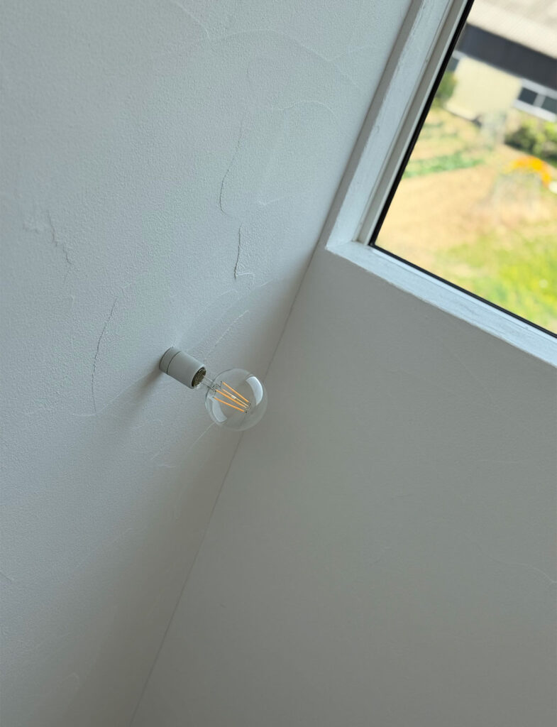 漆喰調の手仕事跡が美しいコテ目仕上げの壁。シンプルな裸電球のブラケットライトと窓からの光が、ニュアンスのある豊かな陰影と素材感を引き立てる壁面のクローズアップです。