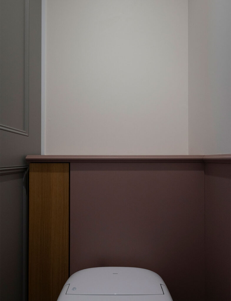 Farrow&Ballの「No.295 Sulking Room Pink」を腰壁に施したトイレインテリア。くすんだ大人のピンクと上部のニュアンスカラーが、スタイリッシュな黒のホルダーや床タイルと調和し、落ち着きのある上品な空間を演出する施工例。