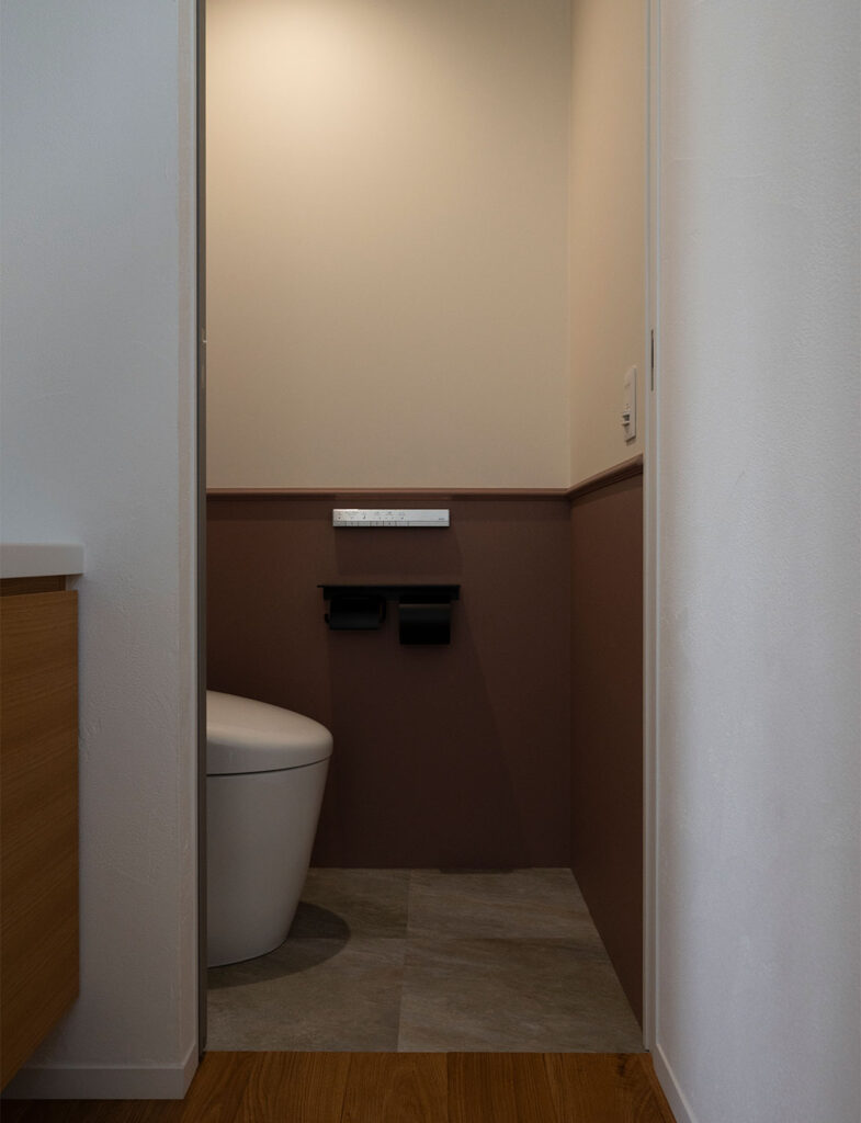 Farrow&Ballの「No.295 Sulking Room Pink」を腰壁に施したトイレインテリア。くすんだ大人のピンクと上部のニュアンスカラーが、スタイリッシュな黒のホルダーや床タイルと調和し、落ち着きのある上品な空間を演出する施工例。