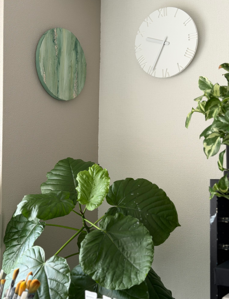 やわらかな壁色を背景に、緑の円形アートや白い時計、花と植物が並ぶ大橋澪さんのアトリエ