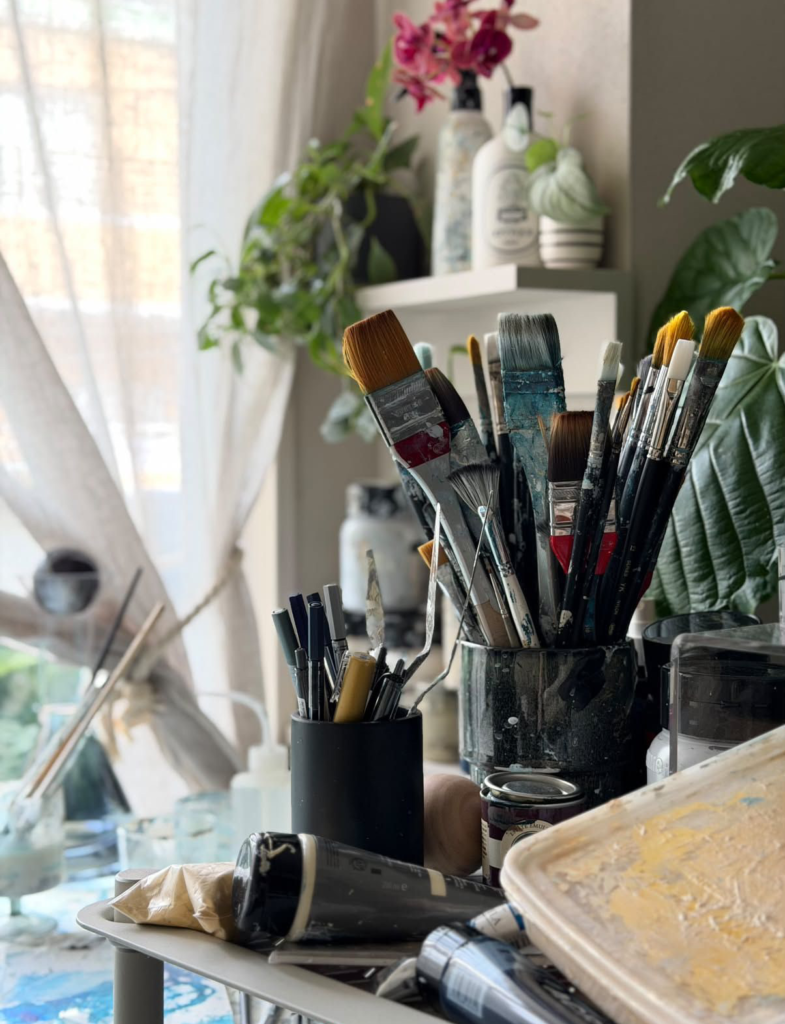 大橋澪さんのアトリエの窓辺に並ぶ、絵筆や刷毛、絵の具の付いた道具。奥には花と植物が飾られている。